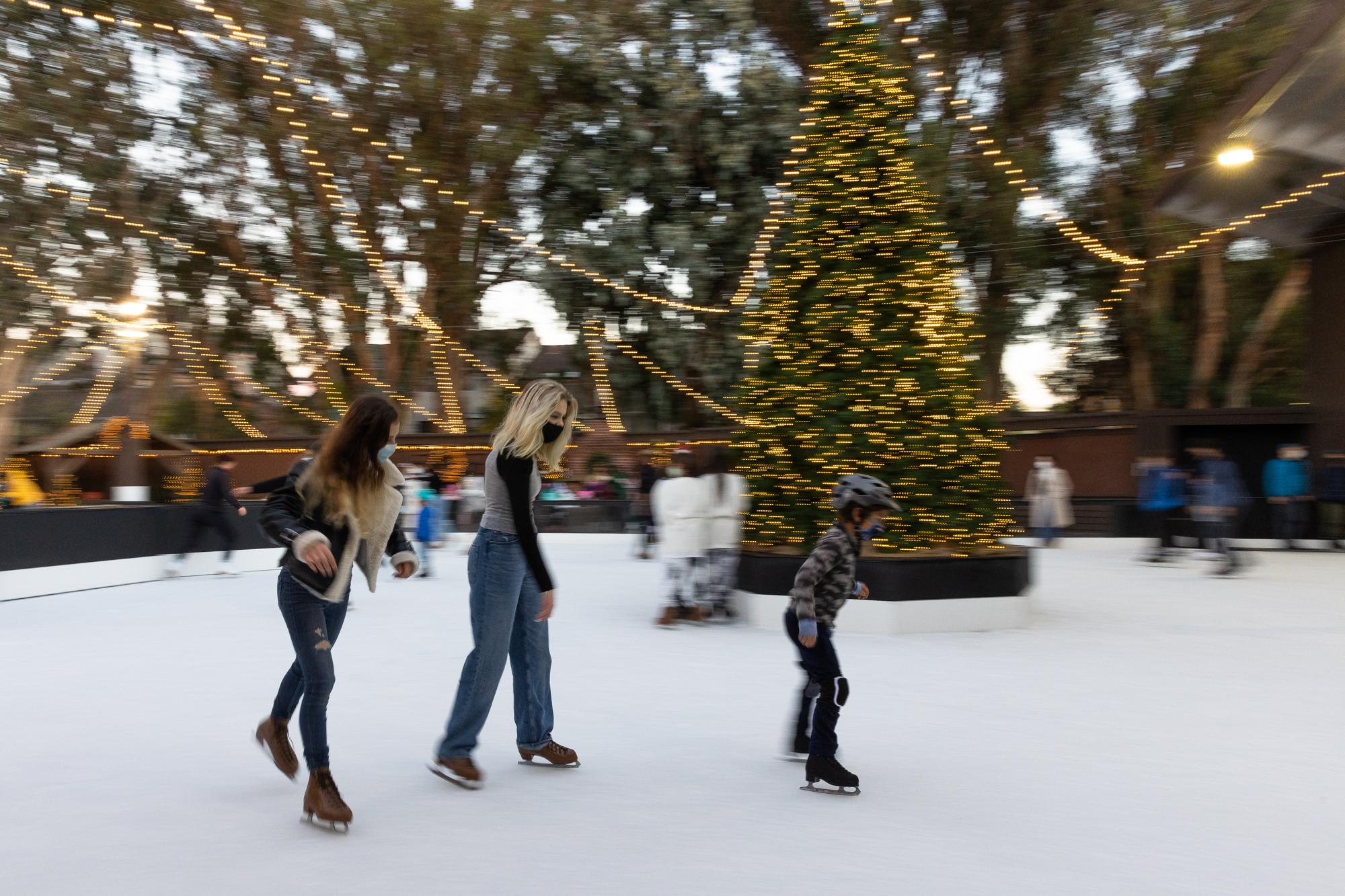 Skaters gliding at dusk beneath a giant lit holiday tree and canopy lights